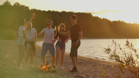 Company-of-boys-and-girls-are-dancing-in-shorts-and-t-shirts-around-bonfire-on-the-sand-beach-with-beer.-They-are-enjoying-the-summer-evening-on-the-river-coast-on-the-open-air-party.